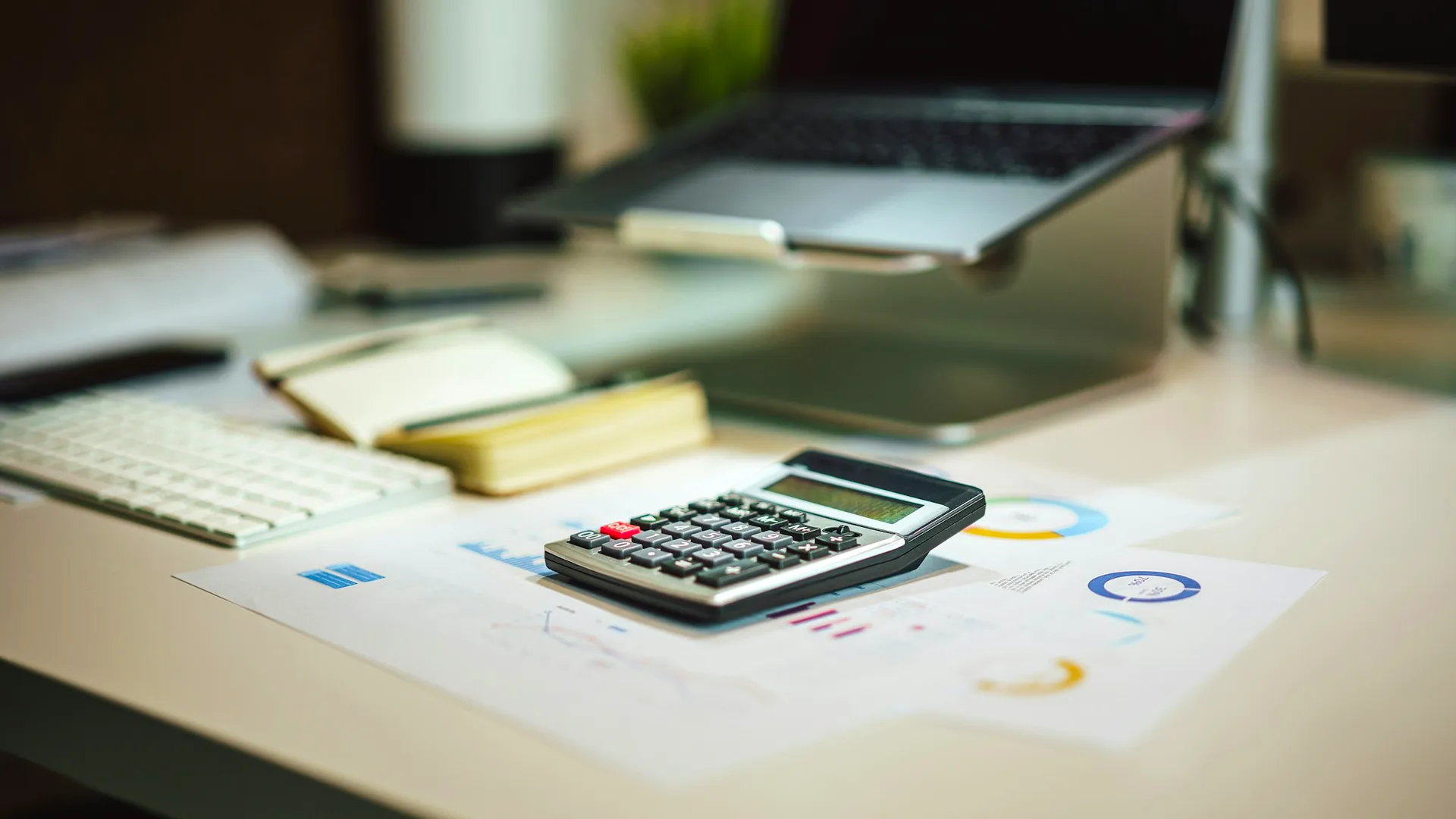 A calculator, reports with charts and a notebook on a bright desk, with a laptop in the background